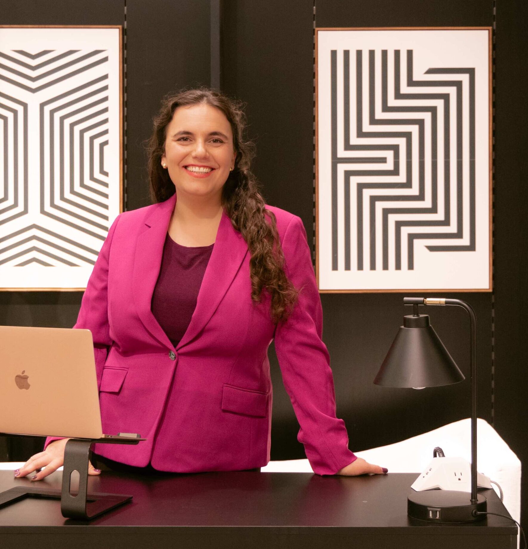 Marketing consultant Rachel Cali is a mid-thirties white woman wearing a pink blazer and standing behind a desk with a laptop on it. She has long, curly brown hair and is smiling directly at the viewer.